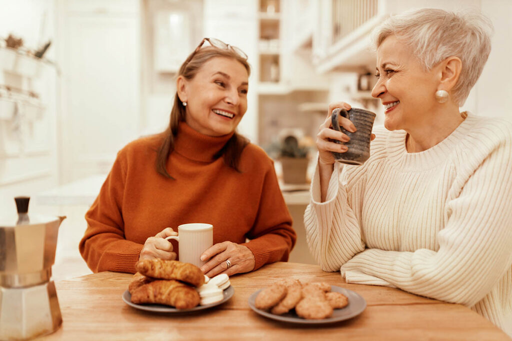 Valley View | Portrait of stylish senior women in cozy sweaters having breakfast together at cafe, sitting at wooden table, sharing news, drinking coffee with pastries