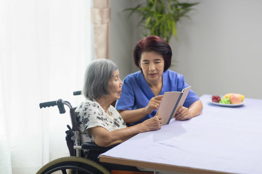 The Ridglea | Senior woman ina wheelchair at the table with her caregiver reading a book